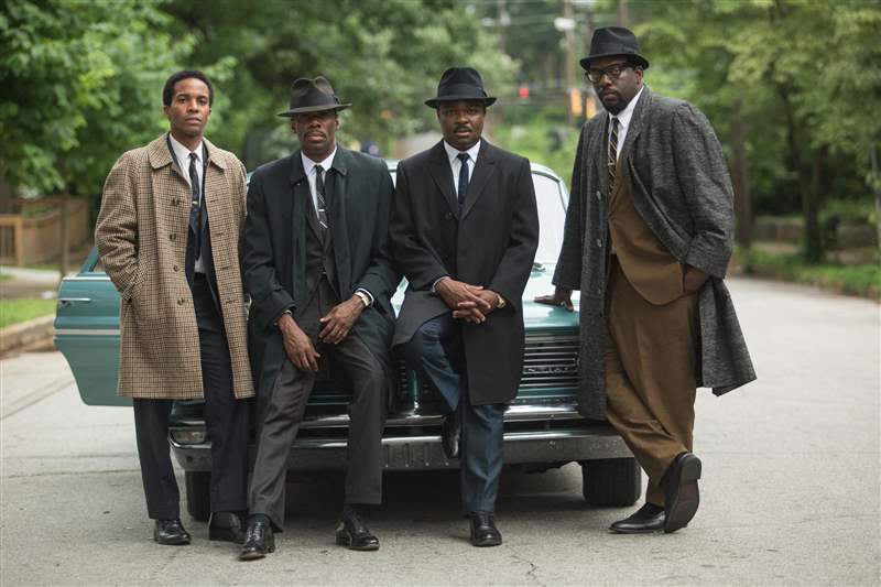 穿著大衣的金 (Oyelowo)、斯泰森 (Stetson)、戴森 (Datejust) 和其他三人在塞爾瑪 (Selma) 的一條街區街道上靠在一輛 1960 年代的汽車上。